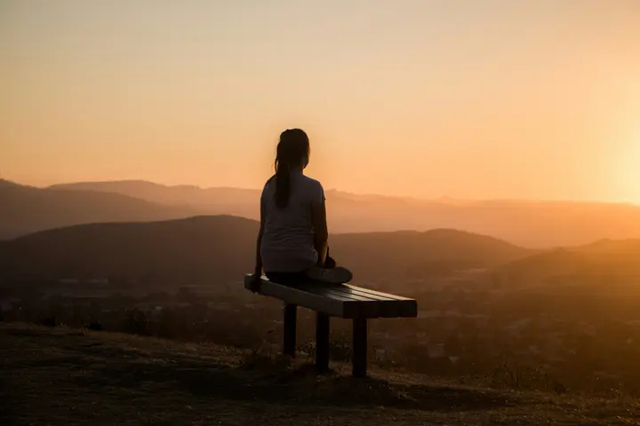 A woman sitting alone on a bench, seen from behind, looking out over hills at sunset.